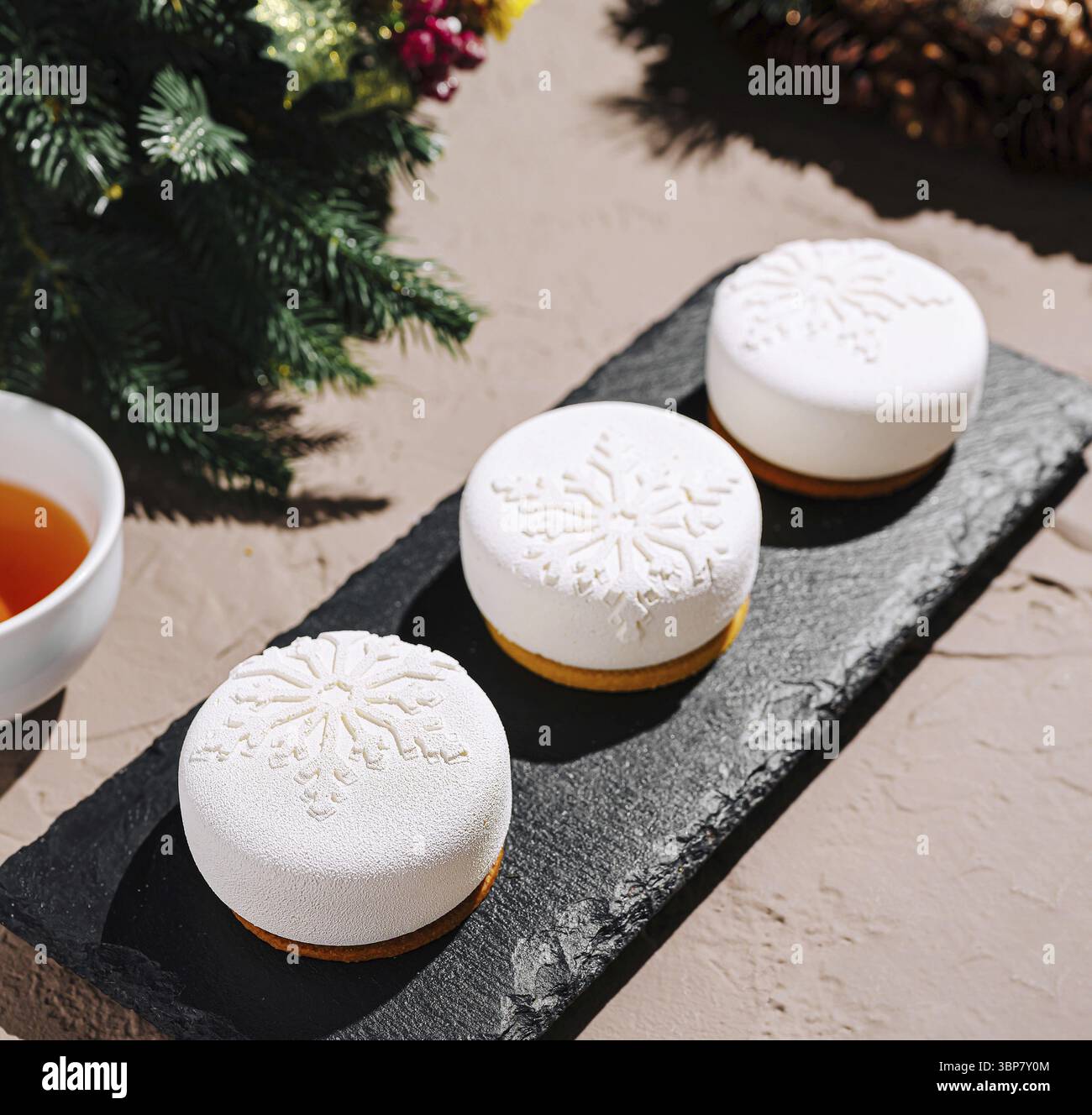 Essen, drei wunderschön gefertigte weiße Desserts mit Schneeflockenmotiven ruhen auf einer schwarzen Schieferplatte, begleitet von einer Tasse Tee. Der festliche dez Stockfoto
