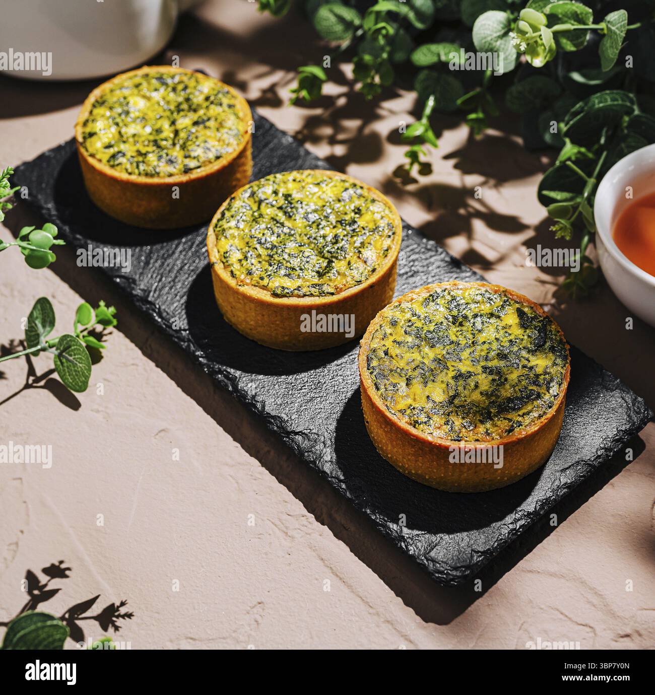 Essen, drei kreisförmige Kräuterquiche sind auf einem dunklen Schiefertablett ausgestellt. Eine Teekanne und eine kleine Schüssel Tee sitzen in der Nähe auf einem rustikalen Tisch, umgeben von Fre Stockfoto