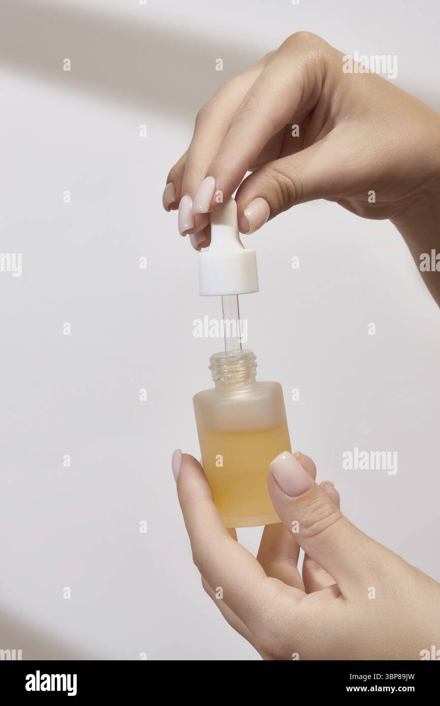 Weibliche Hand mit Tropfer des ätherischen Öls, Aromatherapie-Essenz, Schönheitsserum oder medizinische Flüssigkeit auf weißem Hintergrund. Flaschen ohne Marke für Ihr Desi Stockfoto