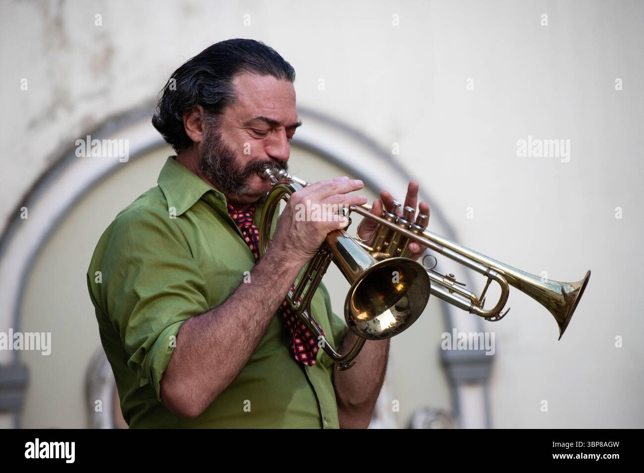 Ein leidenschaftlicher Musiker mit Bart und grünem Hemd spielt eine Messingtrompete mit Stummheit. Stockfoto