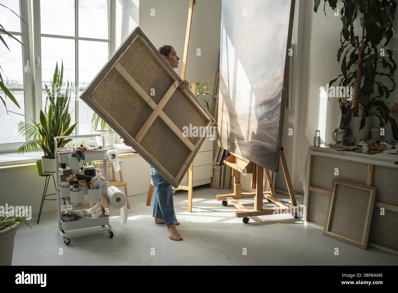 Die interessierte Künstlerin steht mit großer Leinwand in der Hand im Kunststudio und betrachtet die begonnene Malerei auf der Staffelei, auf der Suche nach Inspiration. Kreatives Design Stockfoto