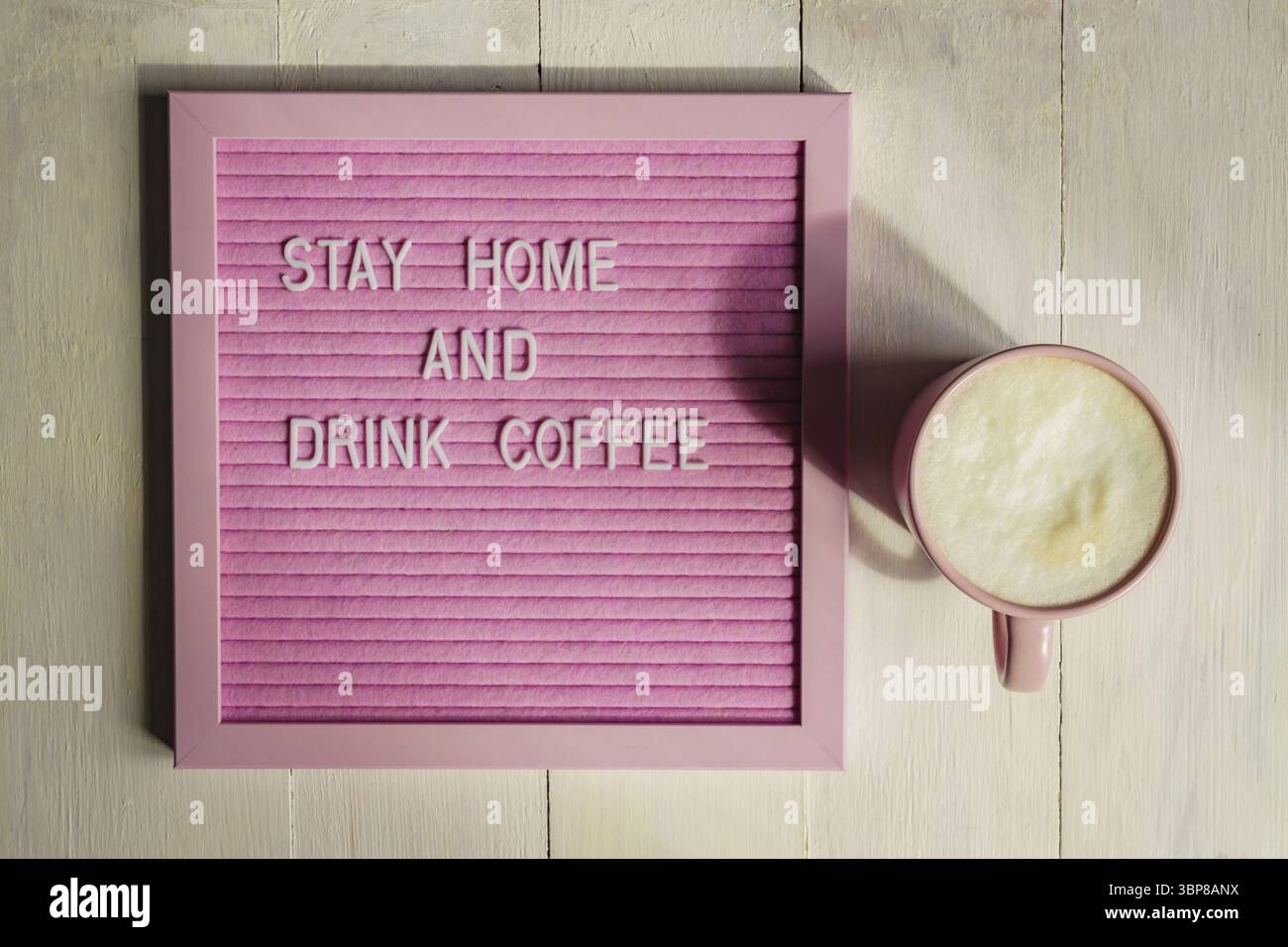 Blick von oben auf Pink Cup mit Cappuccino und Qoute Bleiben Sie zu Hause und trinken Sie Kaffee. Selbstisolierungs- und Quarantänekampagne, um sich während Coronavir zu schützen Stockfoto