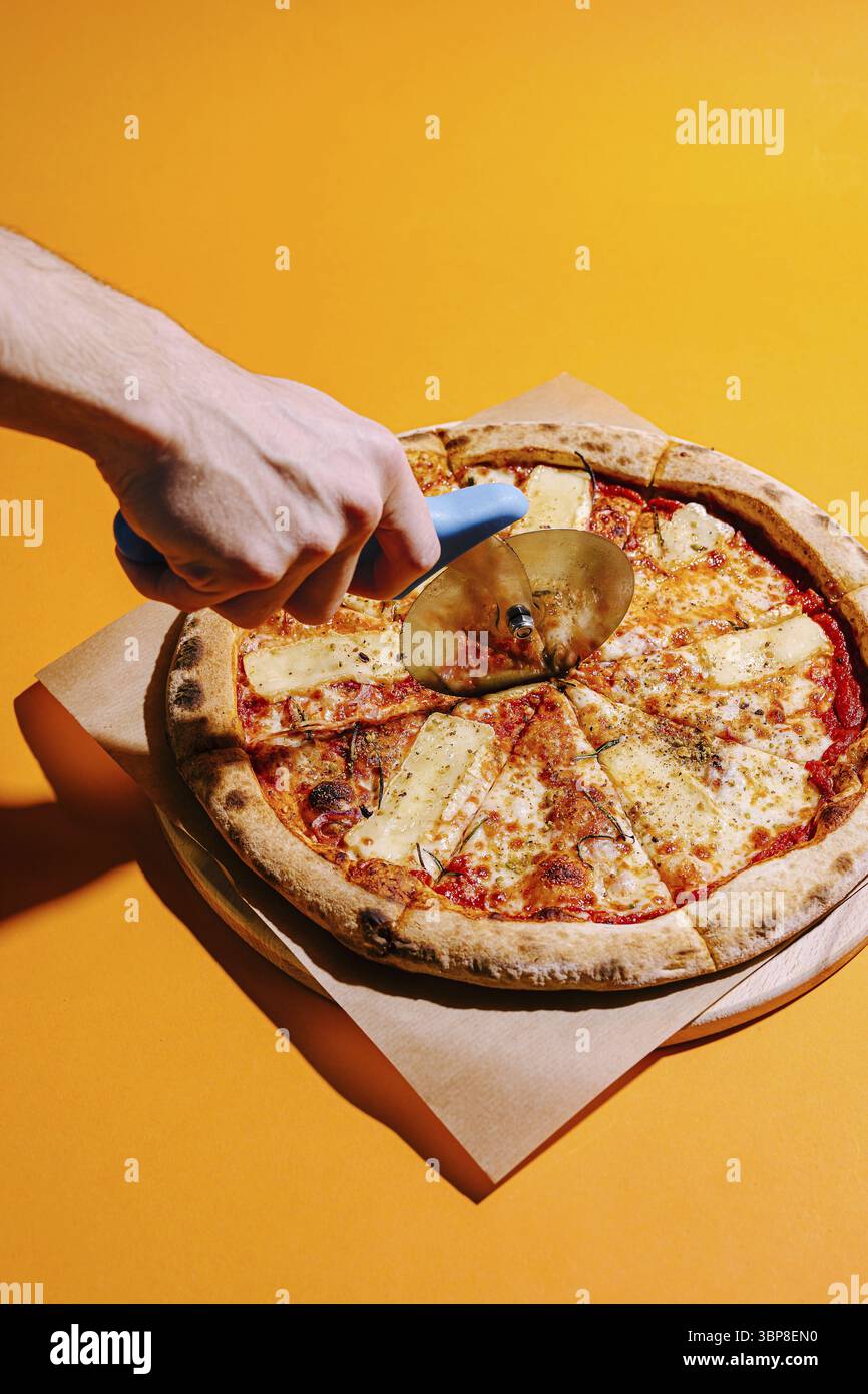 Essen, Eine Hand verwendet einen Pizzaschneider, um eine frische Pizza mit Käse und Ananas zu schneiden. Der hellgelbe Hintergrund verbessert das einladende Erscheinungsbild Stockfoto