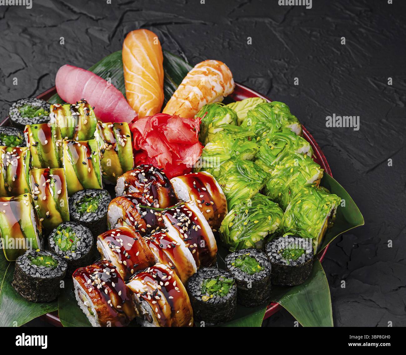 Essen, eine verlockende Platte zeigt eine Vielzahl von Sushi-Brötchen, Sashimi und Gemüsegarnituren, wunderschön angeordnet vor einem strukturierten dunklen Hintergrund Stockfoto