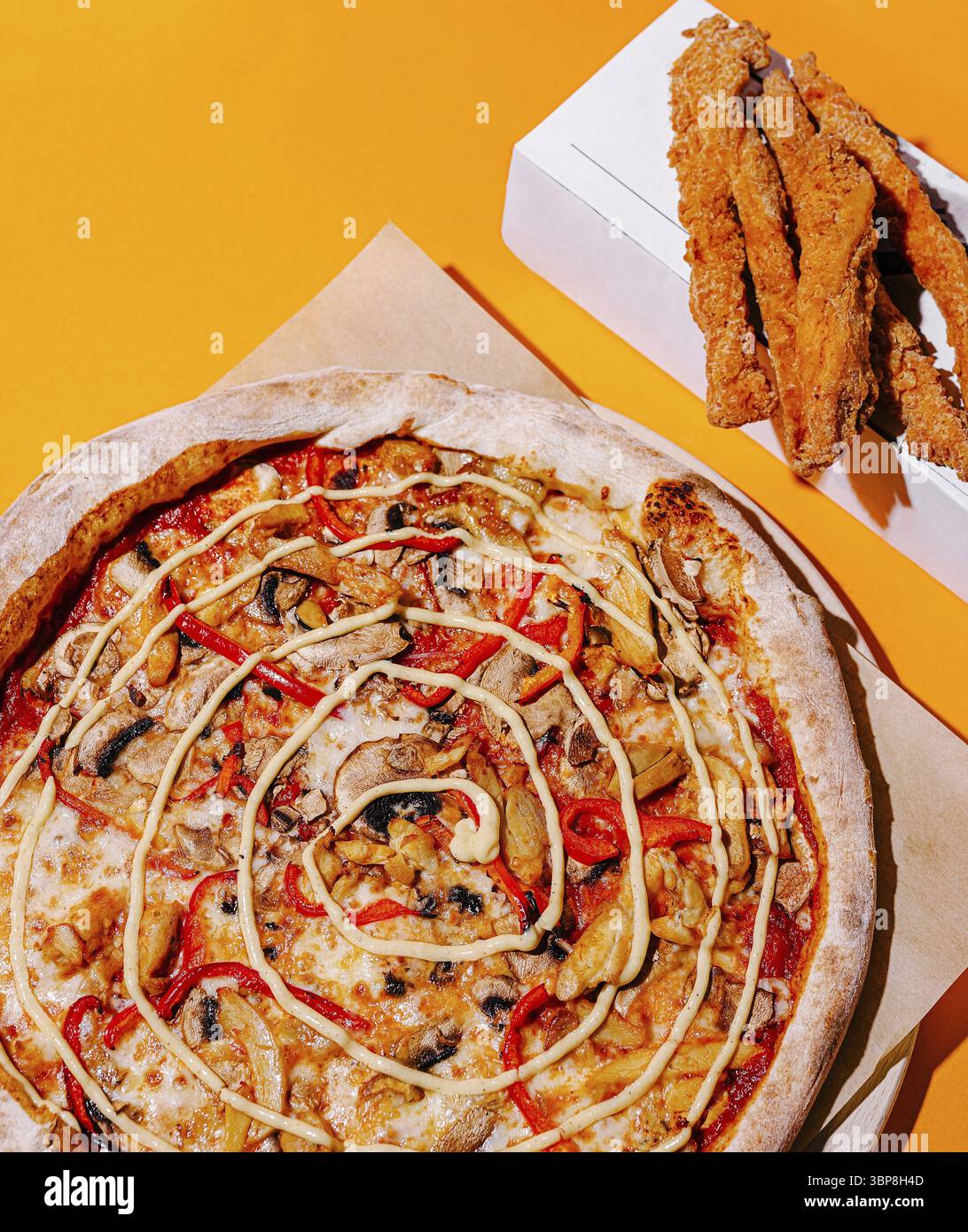Speisen, Eine lebendige Pizza mit Huhn, roter Paprika und einer wirbelnden weißen Sauce wird auf einer hellgelben Oberfläche neben einer Schachtel knusprig gebratener Braten gelegt Stockfoto