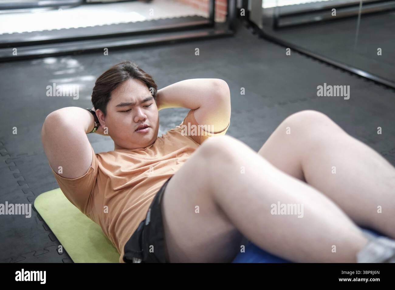 Ein Mann sitzt auf einer Matte. Er trägt ein orangefarbenes Hemd und Shorts. Der Mann schwitzt und hat große Schmerzen Stockfoto