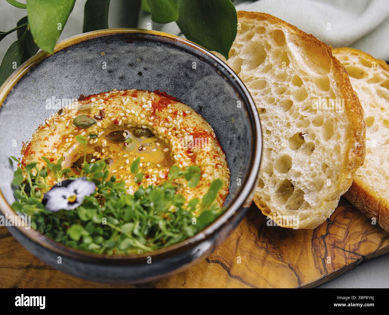 Essen, Schüssel mit cremigem Hummus, garniert mit Samen und einer lebendigen essbaren Blume, serviert mit frischem, handwerklichem Brot Stockfoto