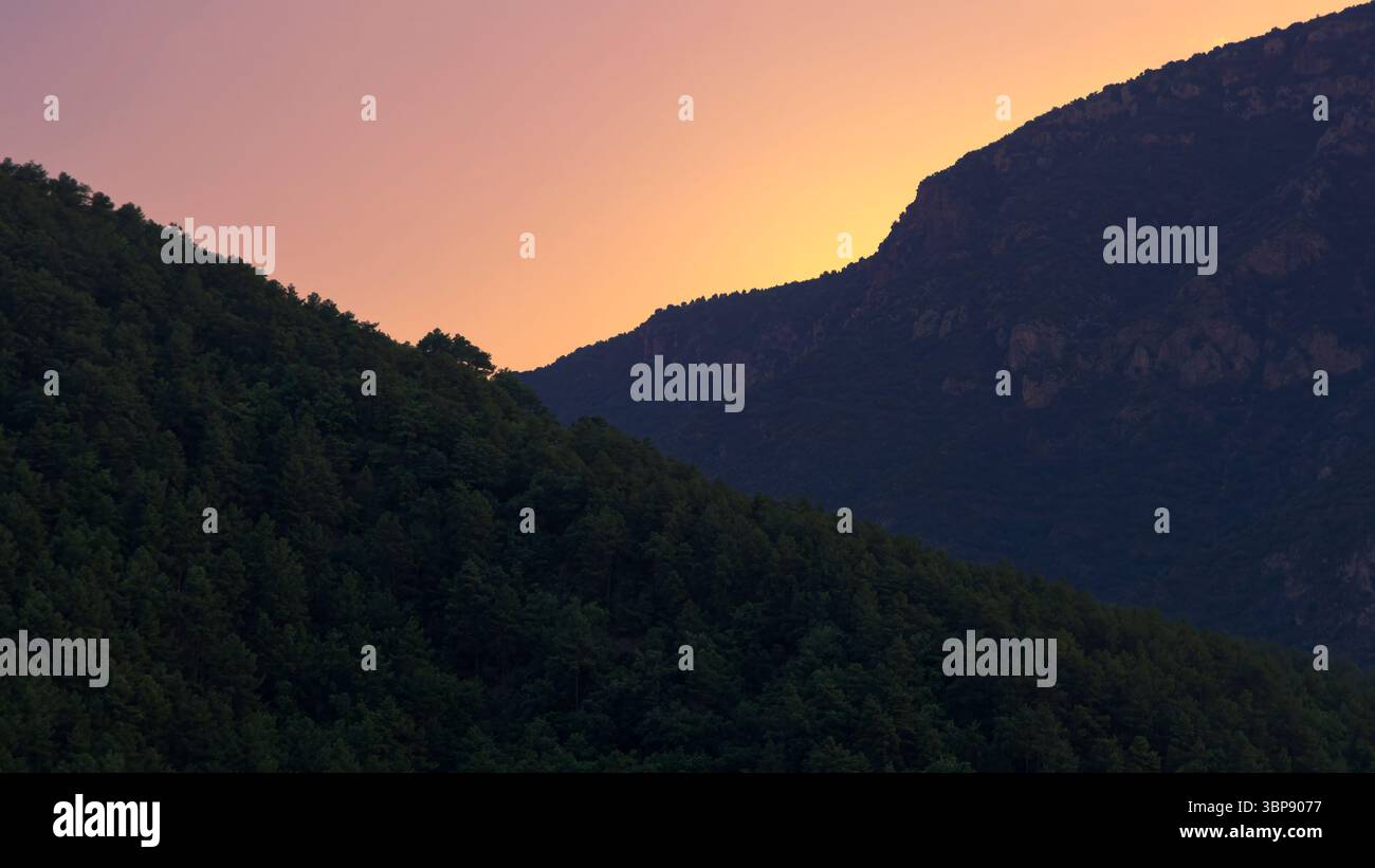 Goldene Farbtöne verschmelzen mit den tiefgrünen Hügeln, während die Sonne hinter den majestätischen Bergen von Alt Urgell in der Dämmerung, Katalonien, untergeht Stockfoto
