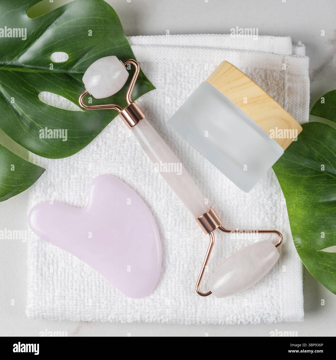 Gesichtsset für die Pflege zu Hause und Spa. Rosenquarz Gesichtswalze, gua Sha Massagegeräte und Glas Gesichtscreme auf weißem Handtuch Hintergrund mit grünem Monstara l Stockfoto