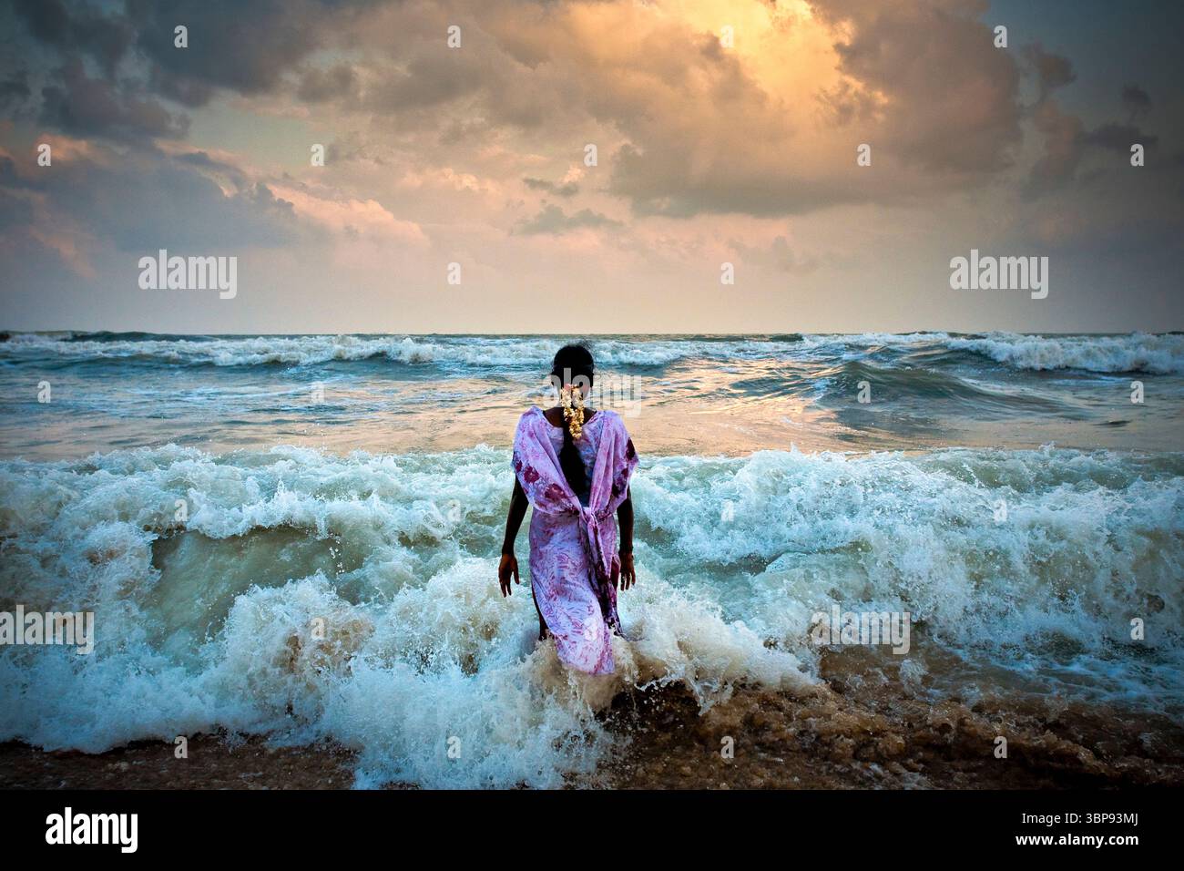 Eine junge Indianerin steht während des Sonnenuntergangs in der Brandung am Strand. Stockfoto