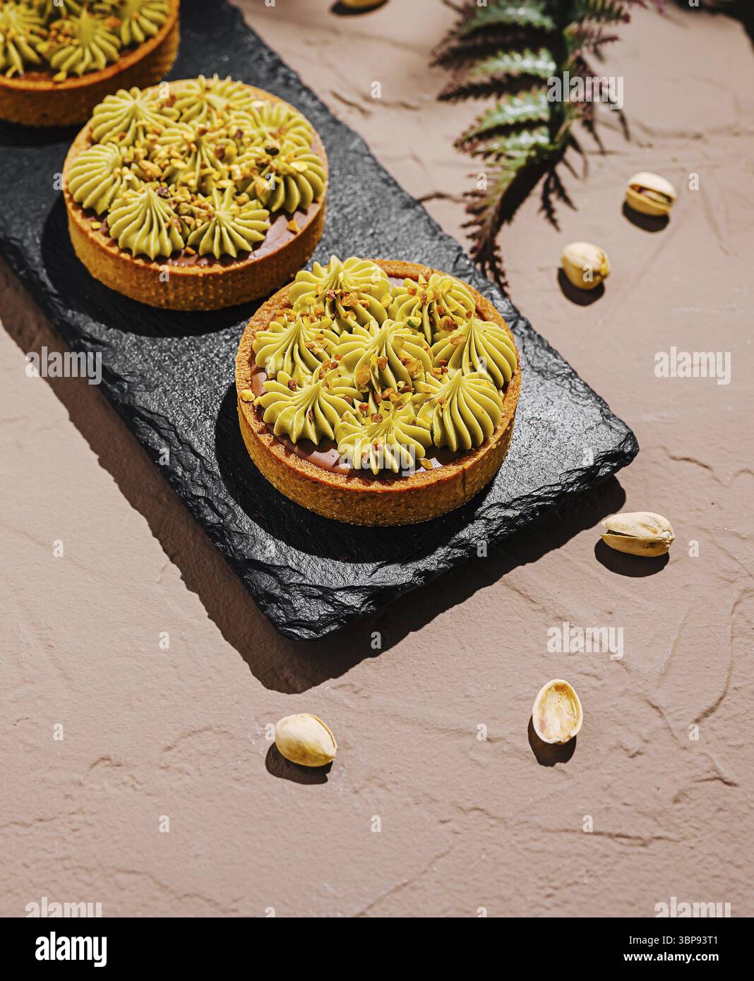 Essen, drei wunderschön zubereitete Pistazientörtchen werden auf einem Schieferbrett ausgestellt. Die Törtchen sind mit leuchtendem grünem Frosting und einer bröckeligen Kruste versehen, Set aga Stockfoto