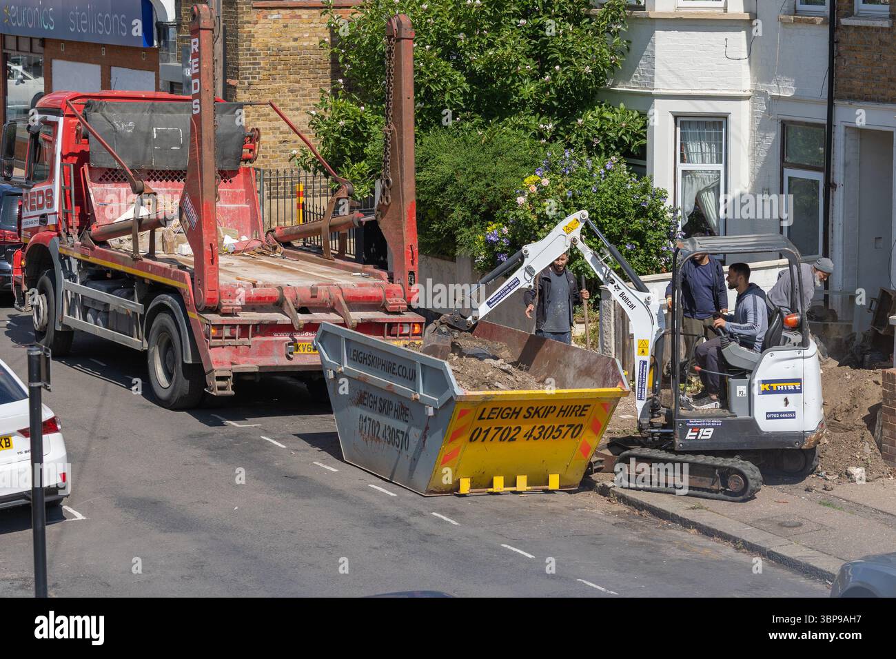 Die Arbeiter beladen einen Sprung mit Abfall mit einem Bobcat E19 und Schaufeln. Stockfoto