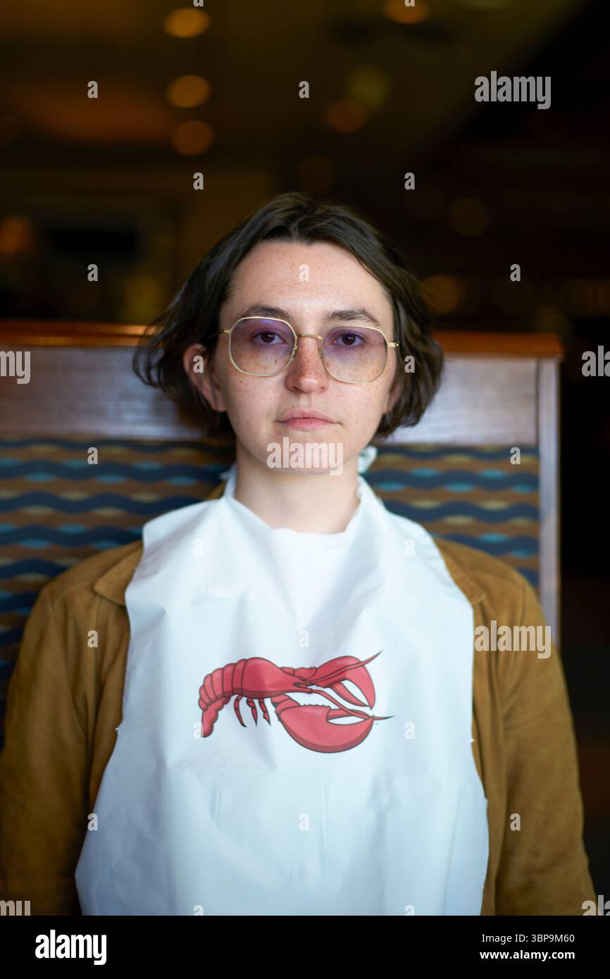 Junge Frau mit Brille und weißem Lätzchen mit rotem Hummermotiv, drinnen mit verschwommenem Hintergrund. Palm Springs, USA Stockfoto