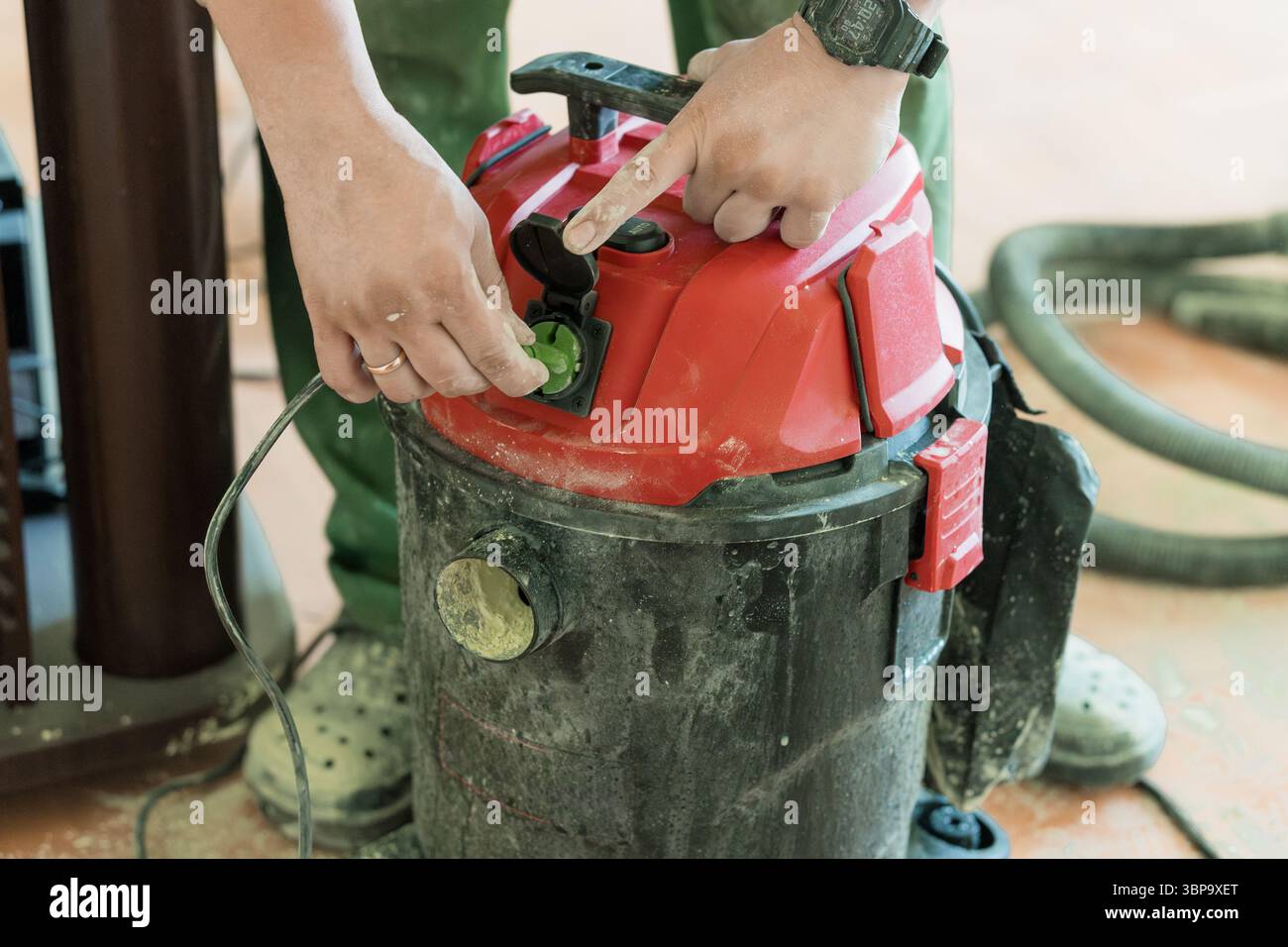 Eine Person in olivgrüner Kleidung verbindet einen grünen Stecker mit einem roten Staubsaugeranschluss in der Werkstatt, was die industrielle Reinigung in einer Werkstatt veranschaulicht Stockfoto