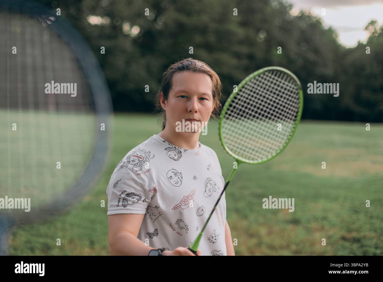 Nahaufnahme Mann mit grünem Badminton Schläger Grasy Park künstlerisches weißes T-Shirt schwarze Shorts Smartwatch neutrales Gesicht natürliches Umgebungslicht Stockfoto