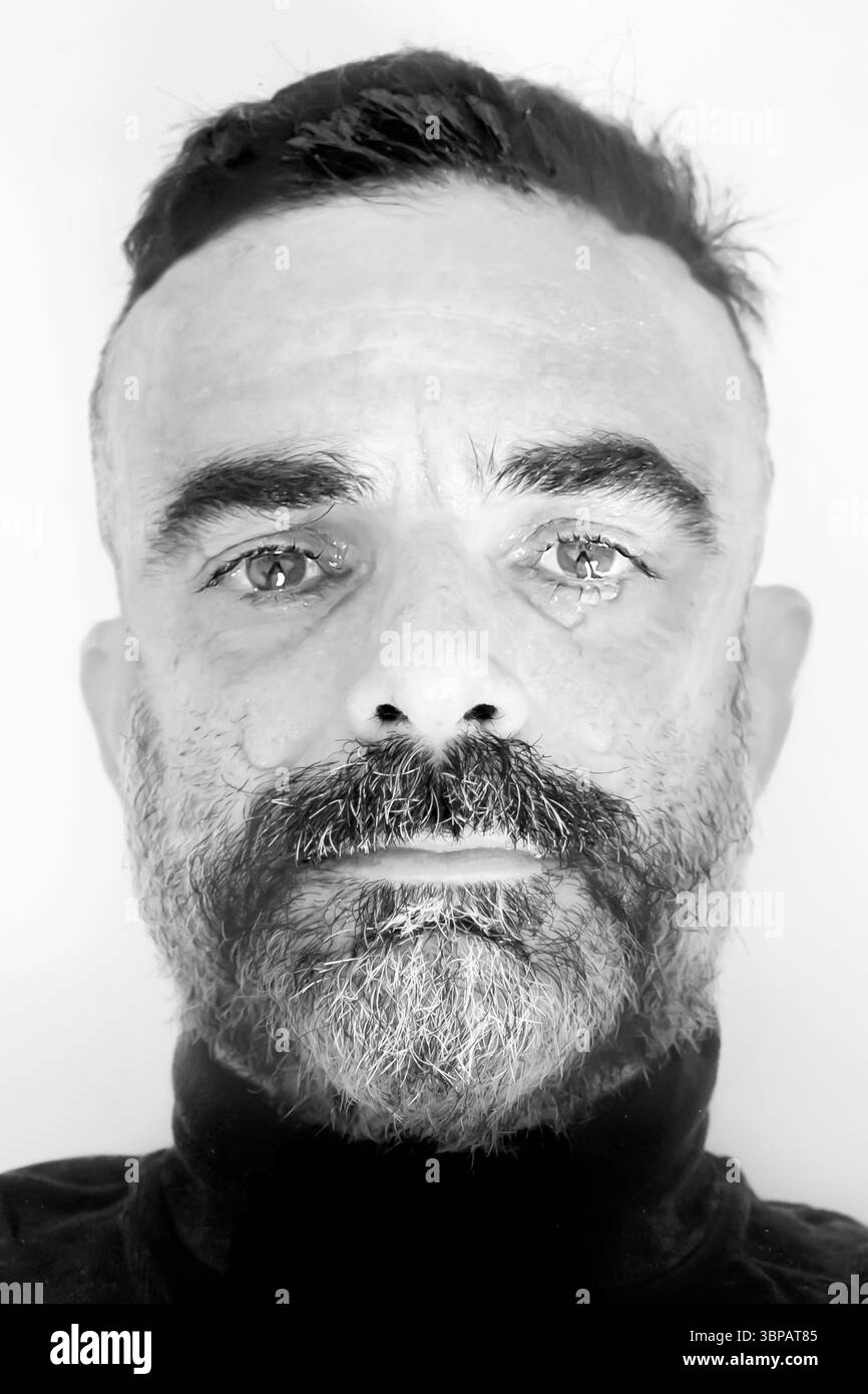Schwarz-weiß-Porträt eines Reifen Mannes mit Bart und kurzen Haaren, der mit ernstem Ausdruck direkt in die Kamera blickt und einen Riss vergießt. Stockfoto