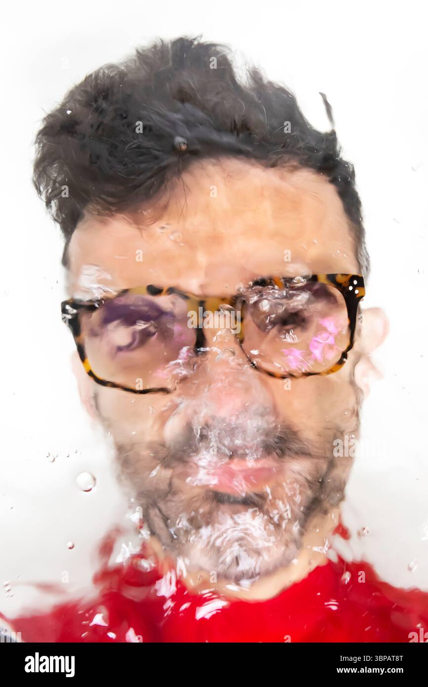 Porträt eines Mannes mit einer Brille durch eine wasserbedeckte Oberfläche, die einen verzerrten und künstlerischen visuellen Effekt mit Blasen und Reflexionen erzeugt. Stockfoto