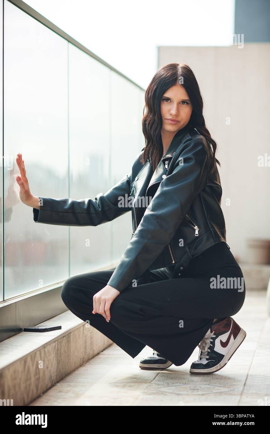 Eine junge Frau, die eine schwarze Lederjacke, eine schwarze Hose und Turnschuhe trägt, hockt sich in der Nähe eines Glasbalkons. Sie hat dunkle Haare und schaut auf die Kamera Stockfoto