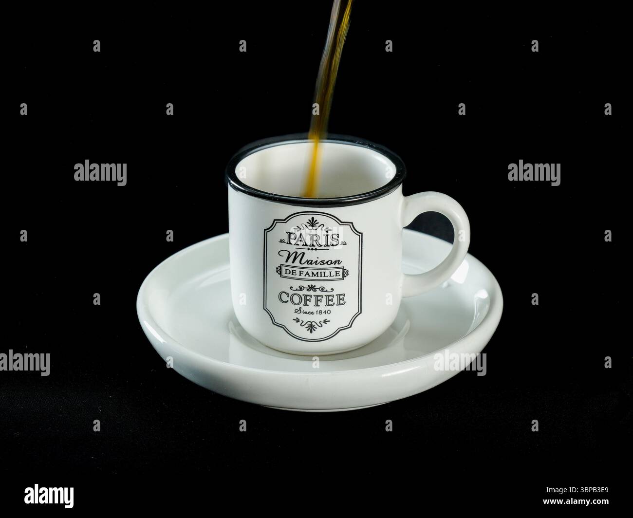 Dunkler Kaffee wird in eine weiße Keramikschale mit einem Vintage-Paris-Design gegossen, die auf einer passenden Untertasse vor einem schwarzen Stoffhintergrund platziert wird. Perfekt. Stockfoto