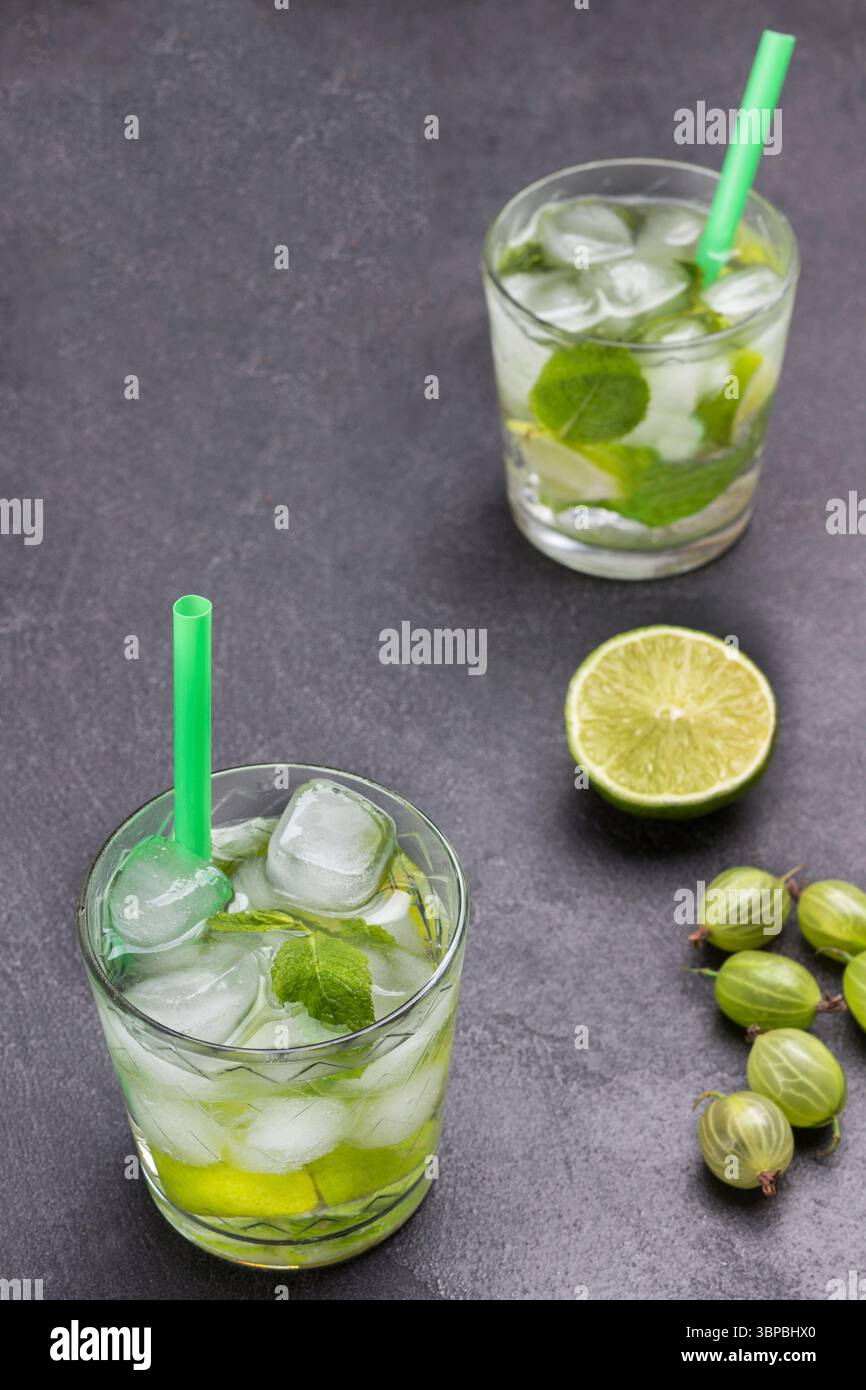 Zwei Gläser kaltes Getränk mit Limette, Minze und Eis. Strohhalm im Glas. Limettenscheiben und Stachelbeeren auf dem Tisch. Schwarzer Hintergrund. Draufsicht Stockfoto