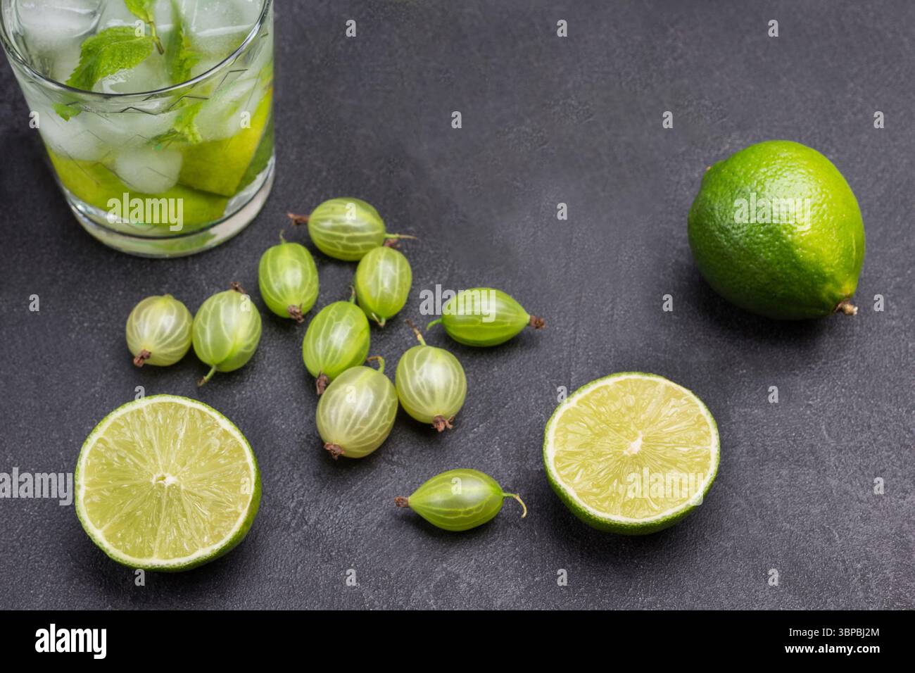 Limettenscheiben und Stachelbeeren auf dem Tisch. Zwei Gläser kaltes Getränk mit Limette, Minze und Eis. Schwarzer Hintergrund. Draufsicht Stockfoto