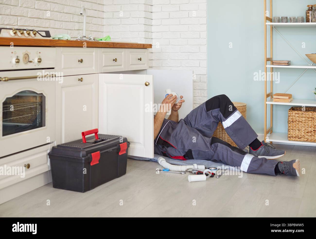 Männlicher Klempner in der Arbeit Uniform auf dem Boden liegen, Küchen-Spülbecken-Rohre befestigen, Sanitär-Service bereitstellen. Stockfoto