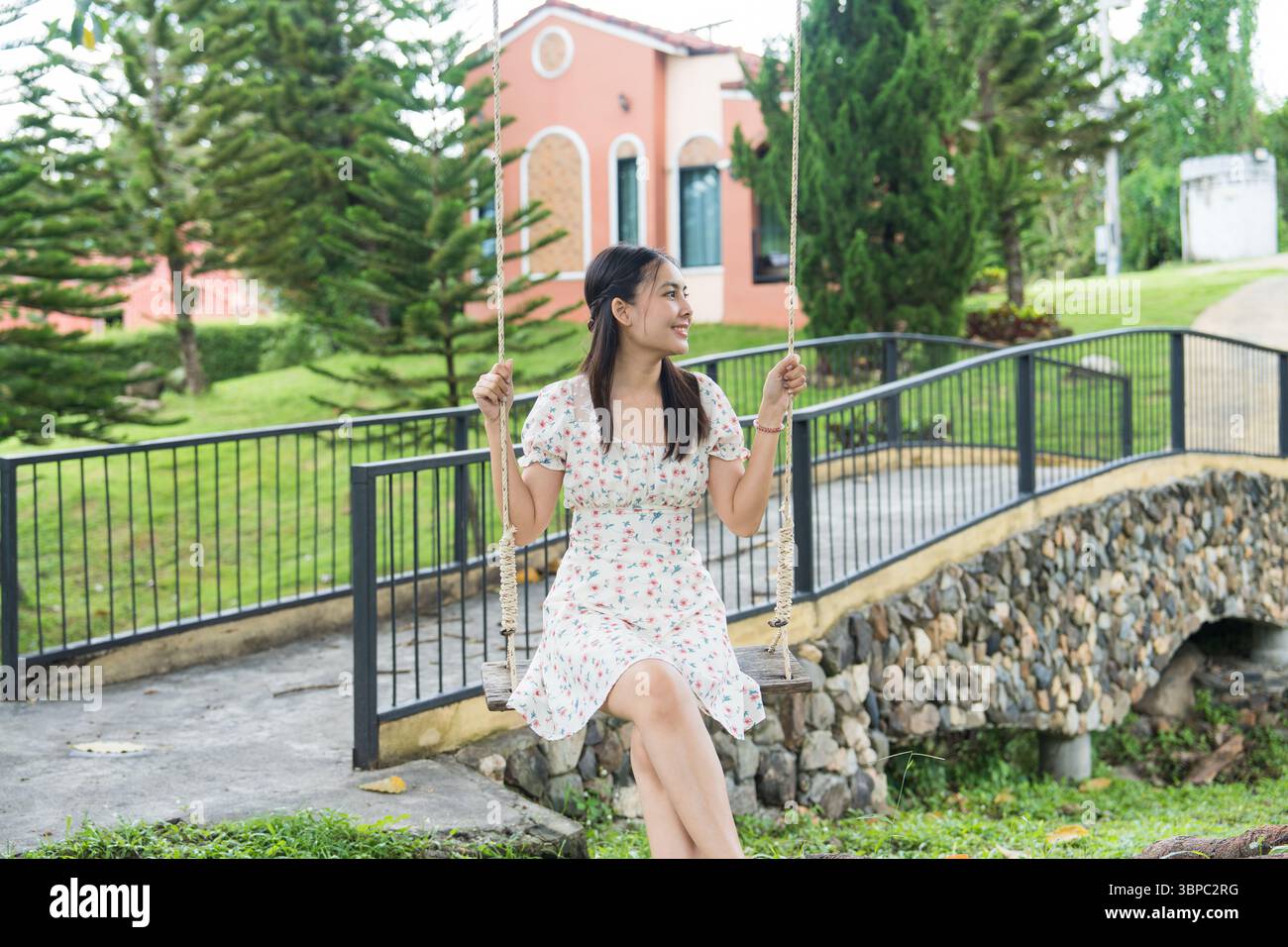 Glückliche asiatische Frau, die auf der Schaukel im tropischen Garten im Resort sitzt und lächelt Stockfoto