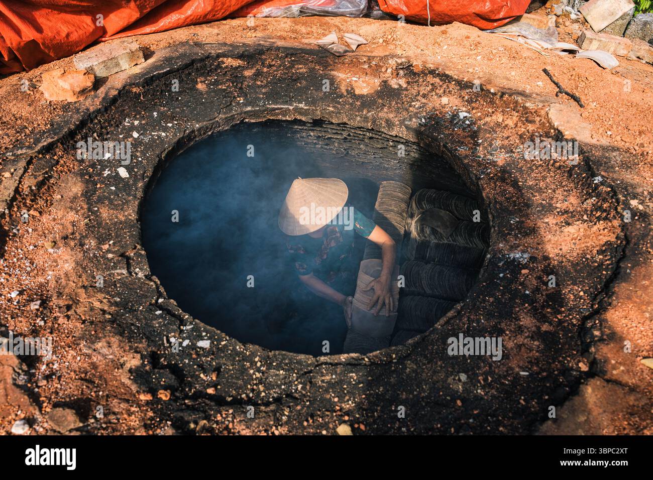 Frau mit Non-la-Hut, die Tonfliesen mit Sonnenlicht in einem rauchigen traditionellen Töpferofen mit ländlicher Handwerkskunst in Vietnam anordnet Stockfoto