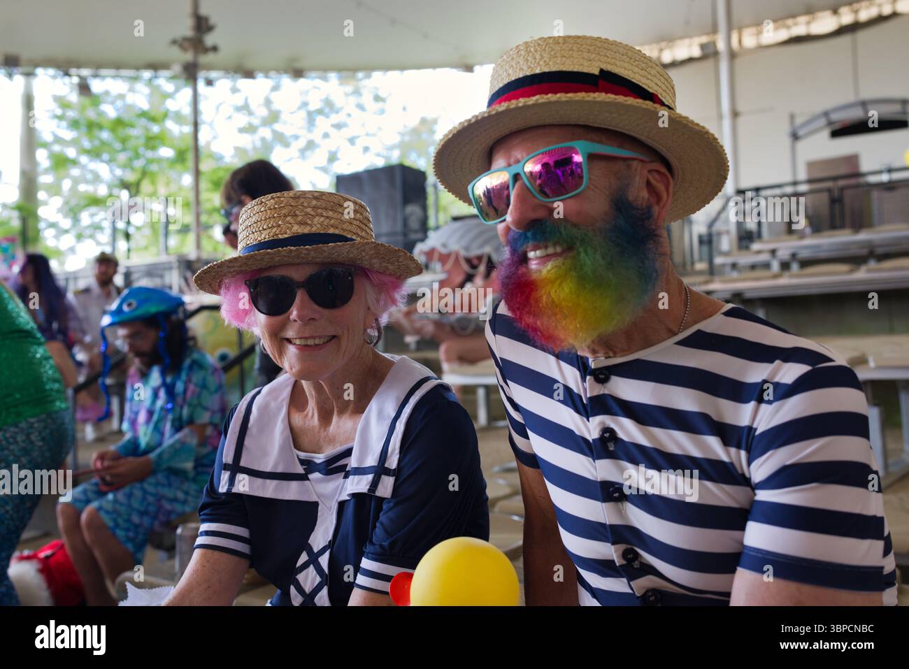 Zwei ältere Erwachsene, ein Mann mit Regenbogenbart und eine Frau mit rosa Haaren, sitzen draußen und tragen Strohhüte und Sonnenbrille. Sie sind in nautischen... Stockfoto