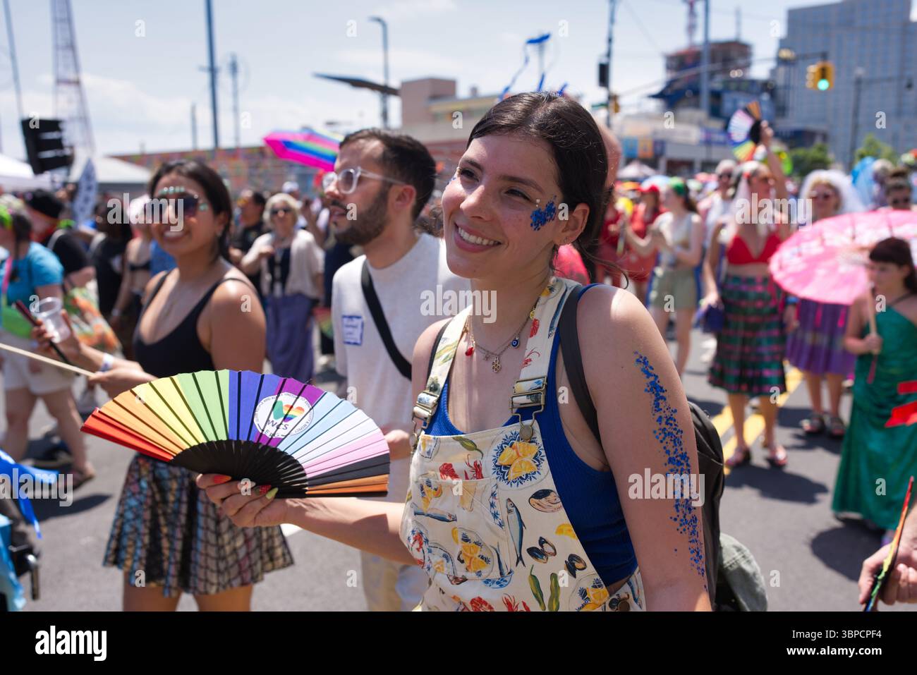 Eine junge Frau mit blauem Glitzer an Arm und Gesicht hält einen Regenbogen-Handfan bei einem lebhaften Outdoor-Event. Sie trägt einen gemusterten Jumpsuit. Andere in Stockfoto