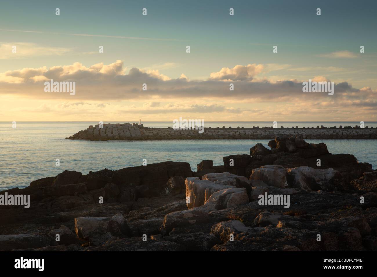 Sanftes Sonnenlicht taucht bei Sonnenaufgang eine ruhige Meereslandschaft auf und hebt die felsige Küste und den Wellenbrecher in Ericeira, Portugal, hervor Stockfoto