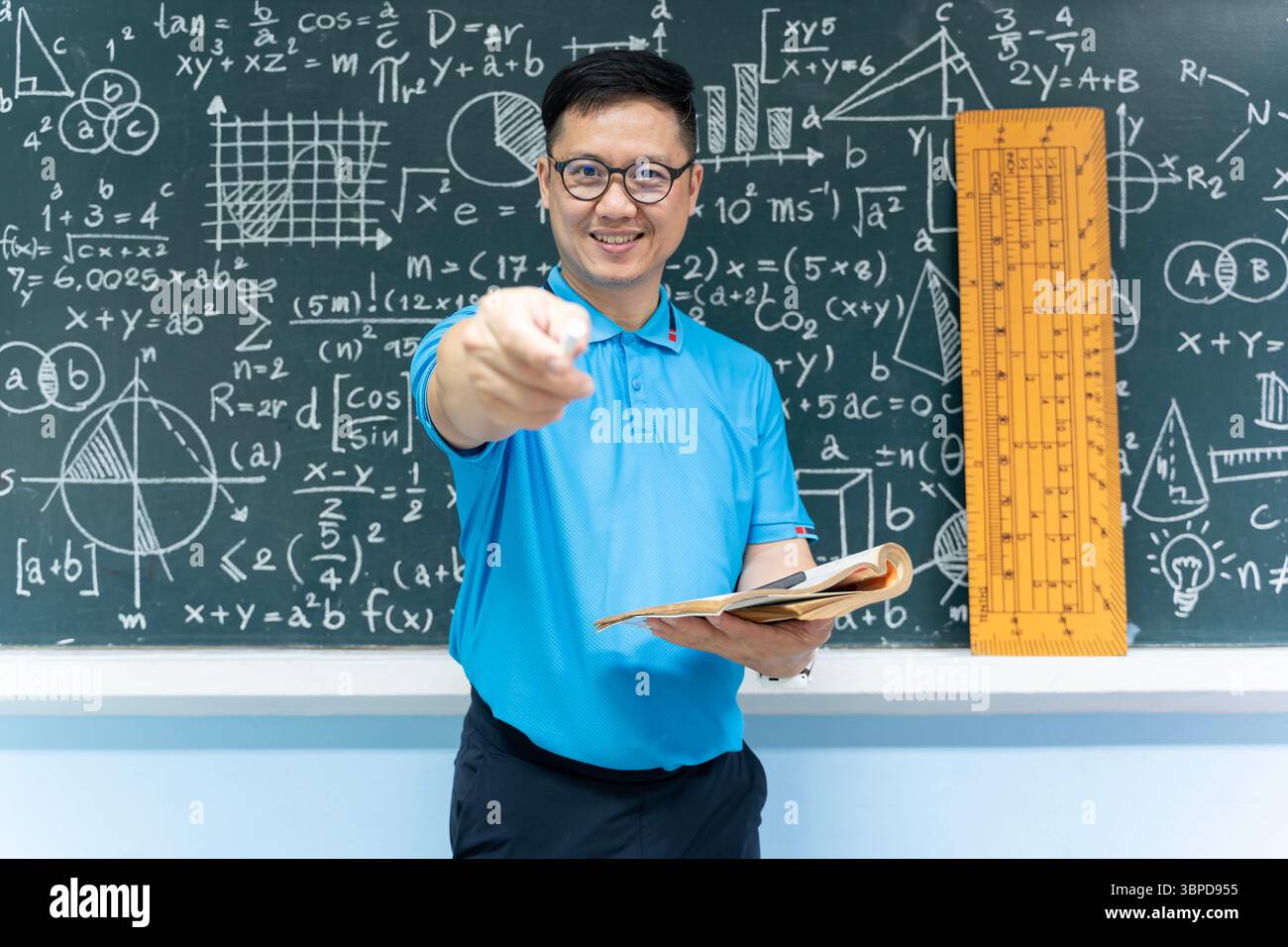 Asiatischer Mathematiklehrer in blauem Hemd lächelt und zeigt weiße Kreide in Richtung Kamera, während er das Buch mit Mathe formul vor der Tafel hält Stockfoto