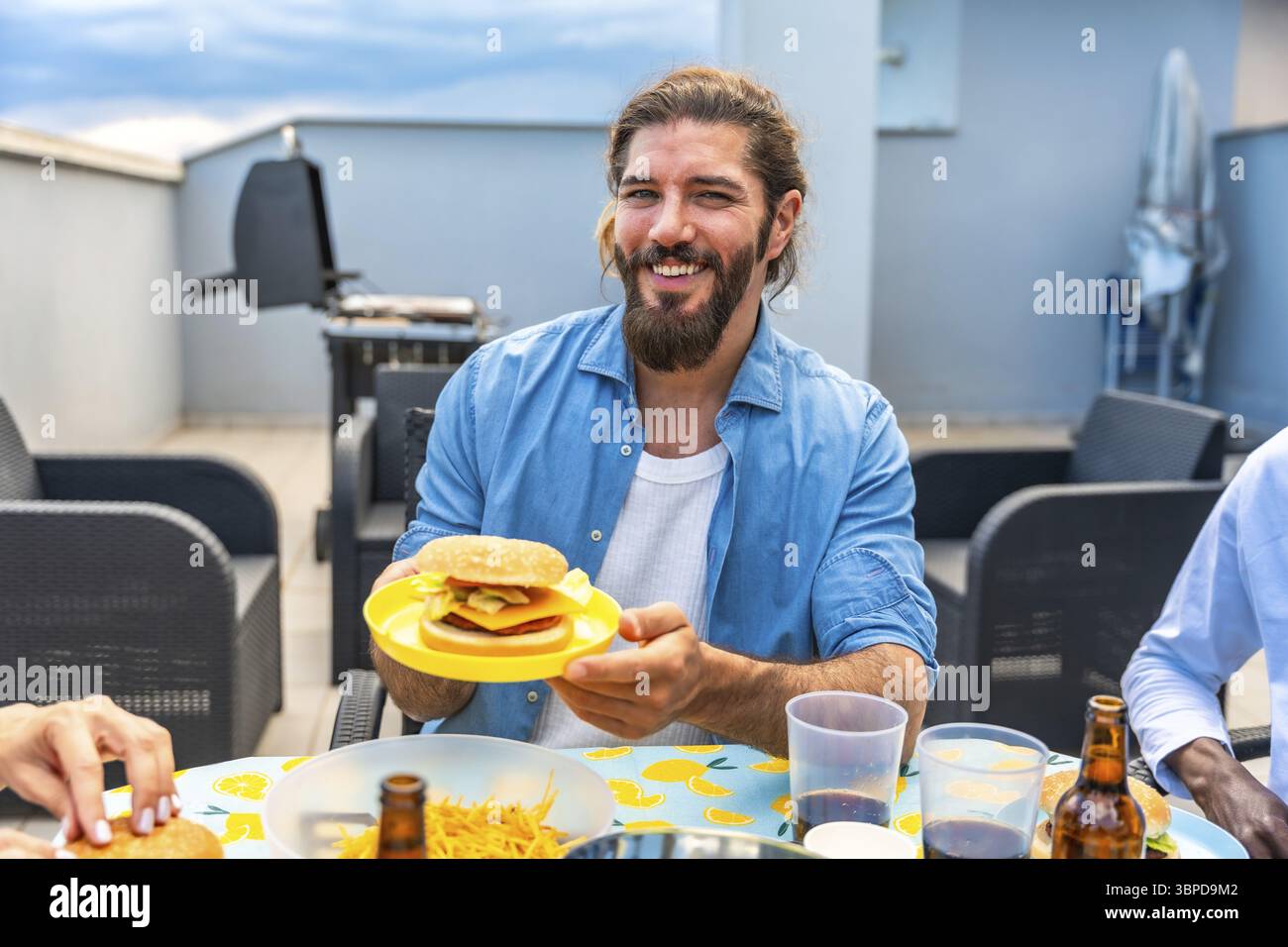 Ein glücklicher Mann hält einen Burger während einer Grillparty auf dem Dach mit Freunden und genießt gutes Essen und Gesellschaft Stockfoto