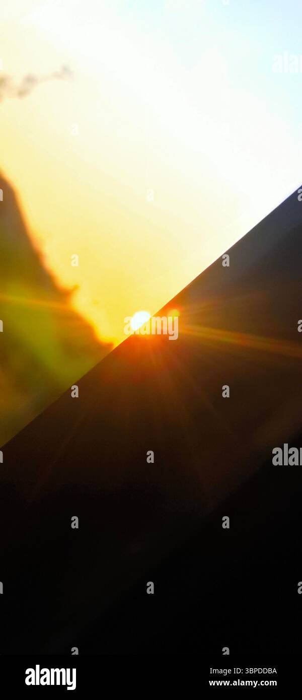 Kreativer Blick auf die untergehende Sonne, die teilweise durch ein diagonales Glas blockiert wird und bei Sonnenuntergang warme Sonnenstrahlen durch den goldenen Himmel wirft. Stockfoto