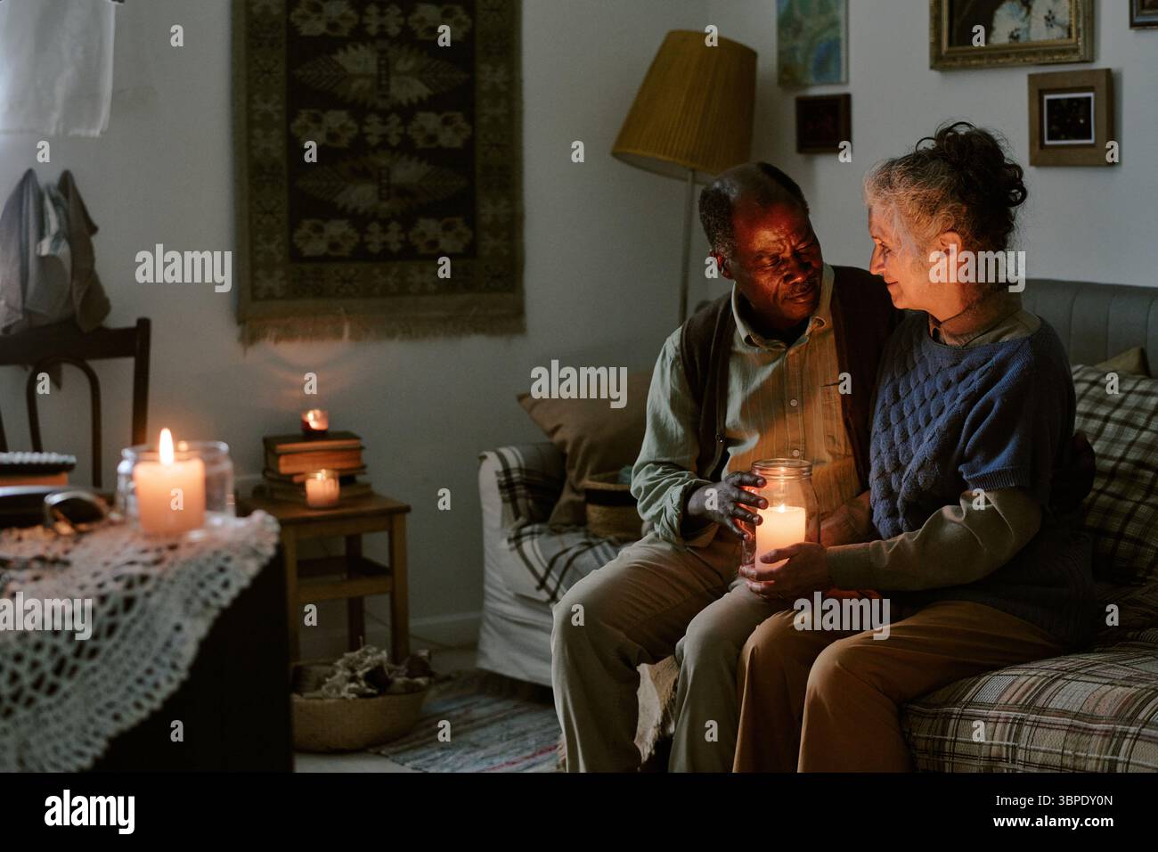 Ältere multiethnische Paare sitzen zusammen und halten Kerze in einem bescheidenen Wohnzimmer Stockfoto