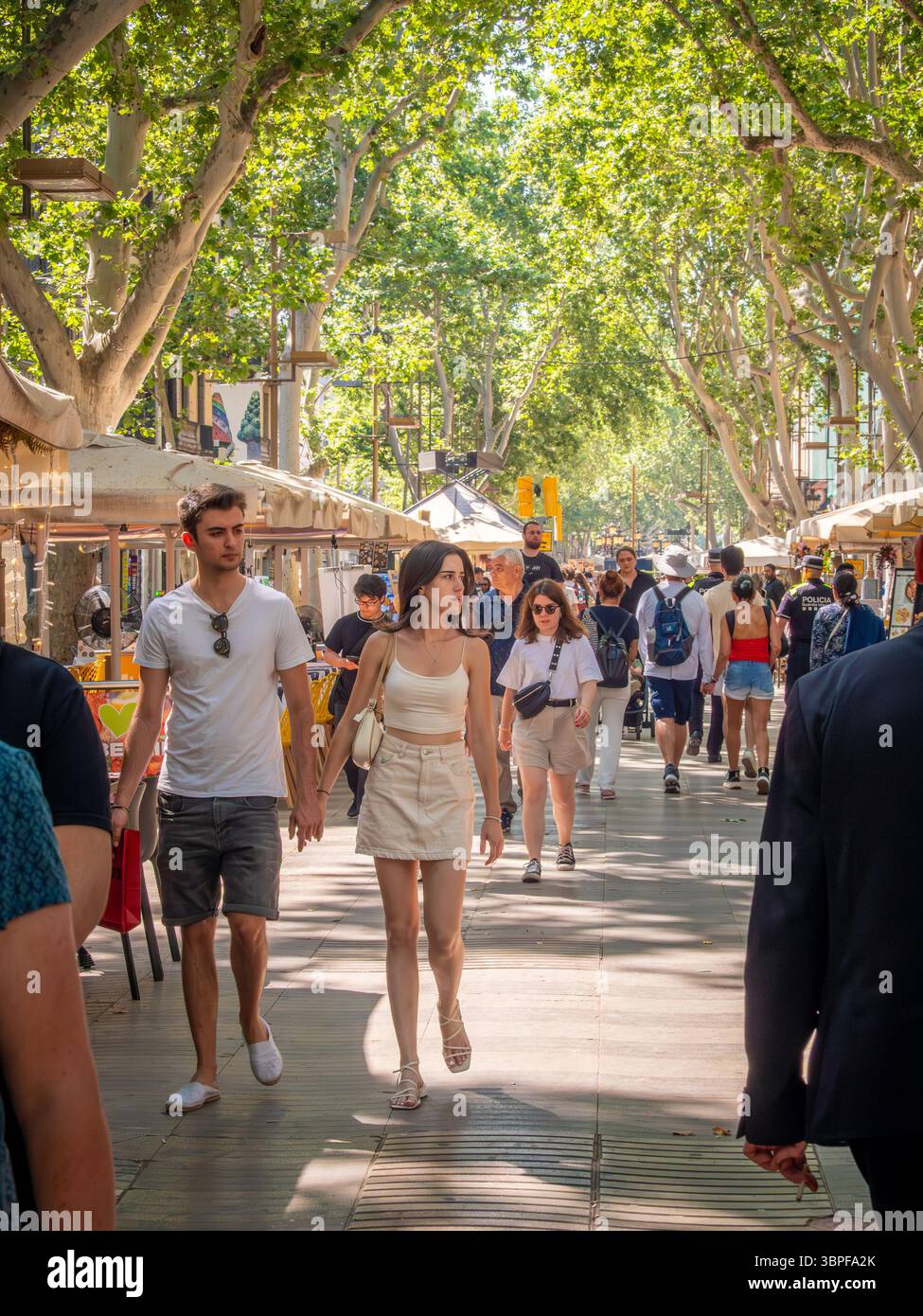 Ein junges Paar hält Hände, während es mit Menschenmassen auf der berühmten, von Bäumen gesäumten Promenade La Rambla in Barcelona, Spanien, spaziert. Stockfoto