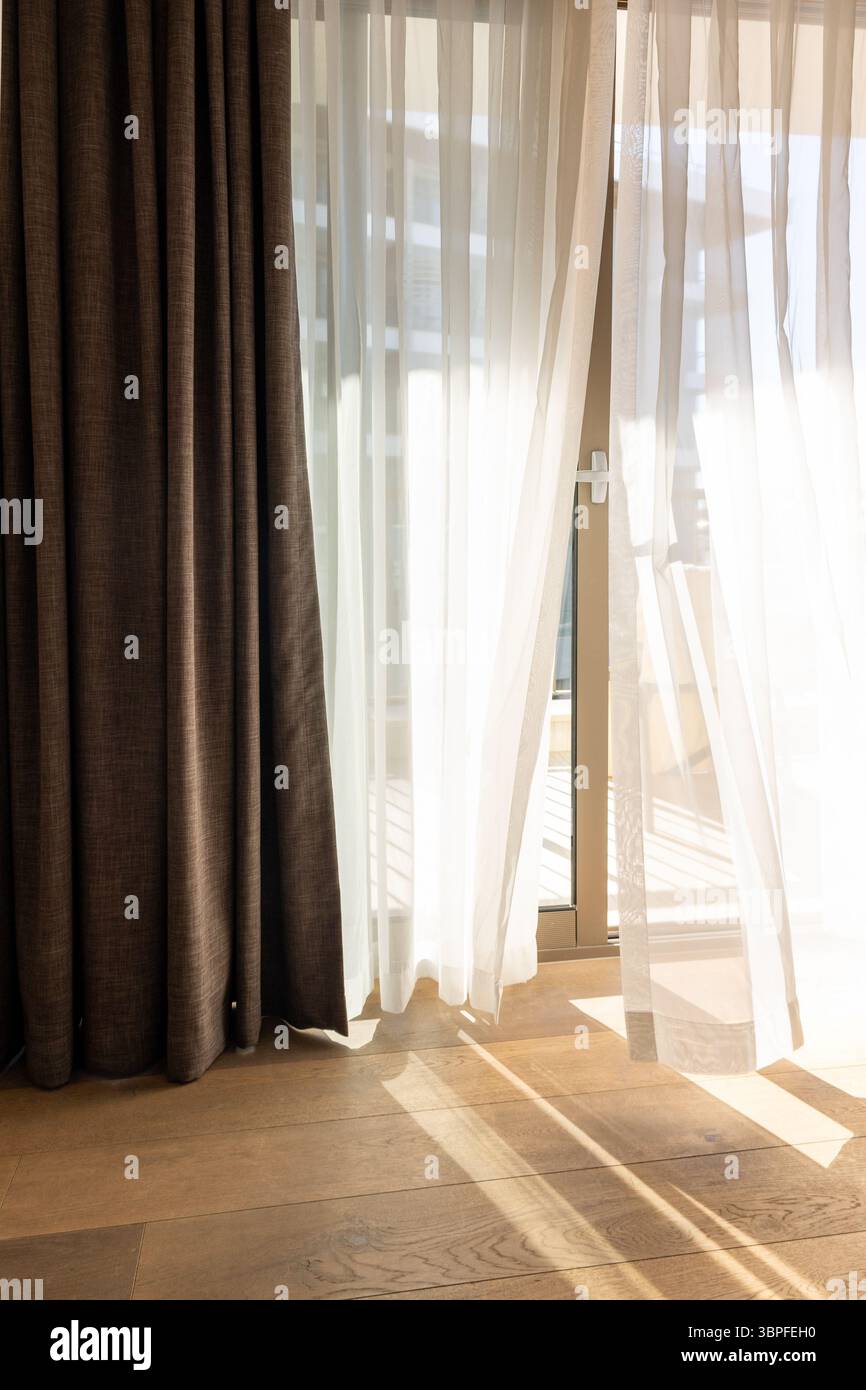 Moderne Zimmereinrichtung mit offenem Fenster in minimalem Stil. Transparenter Tüll mit morgendlichen Sonnenstrahlen auf Schattenhintergrund. Vertikales Foto. Stockfoto