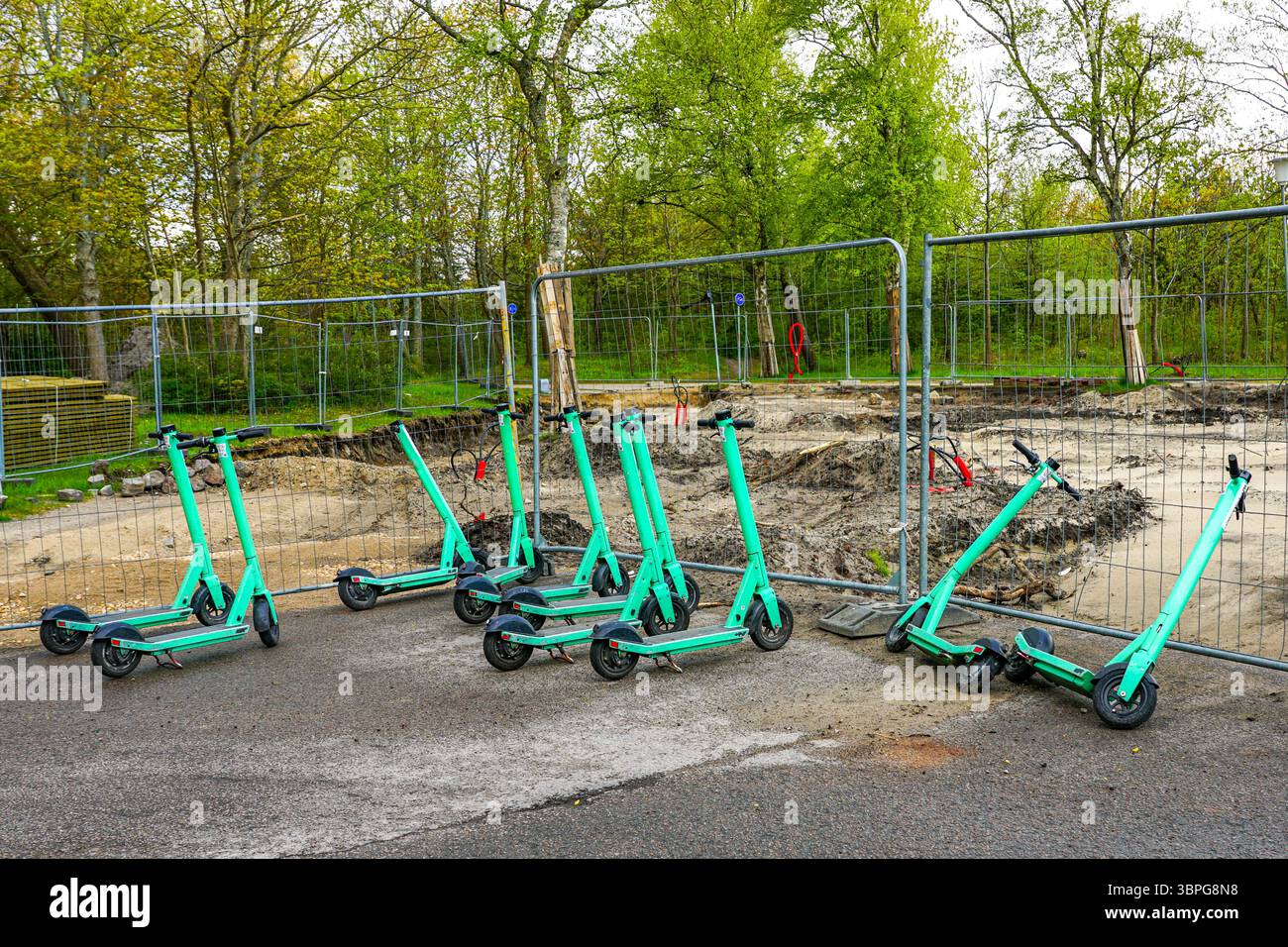 Gemeinsam genutzte Elektroroller, die auf einer Baustelle mit Sicherheitszaun und Baumhintergrund geparkt sind Stockfoto