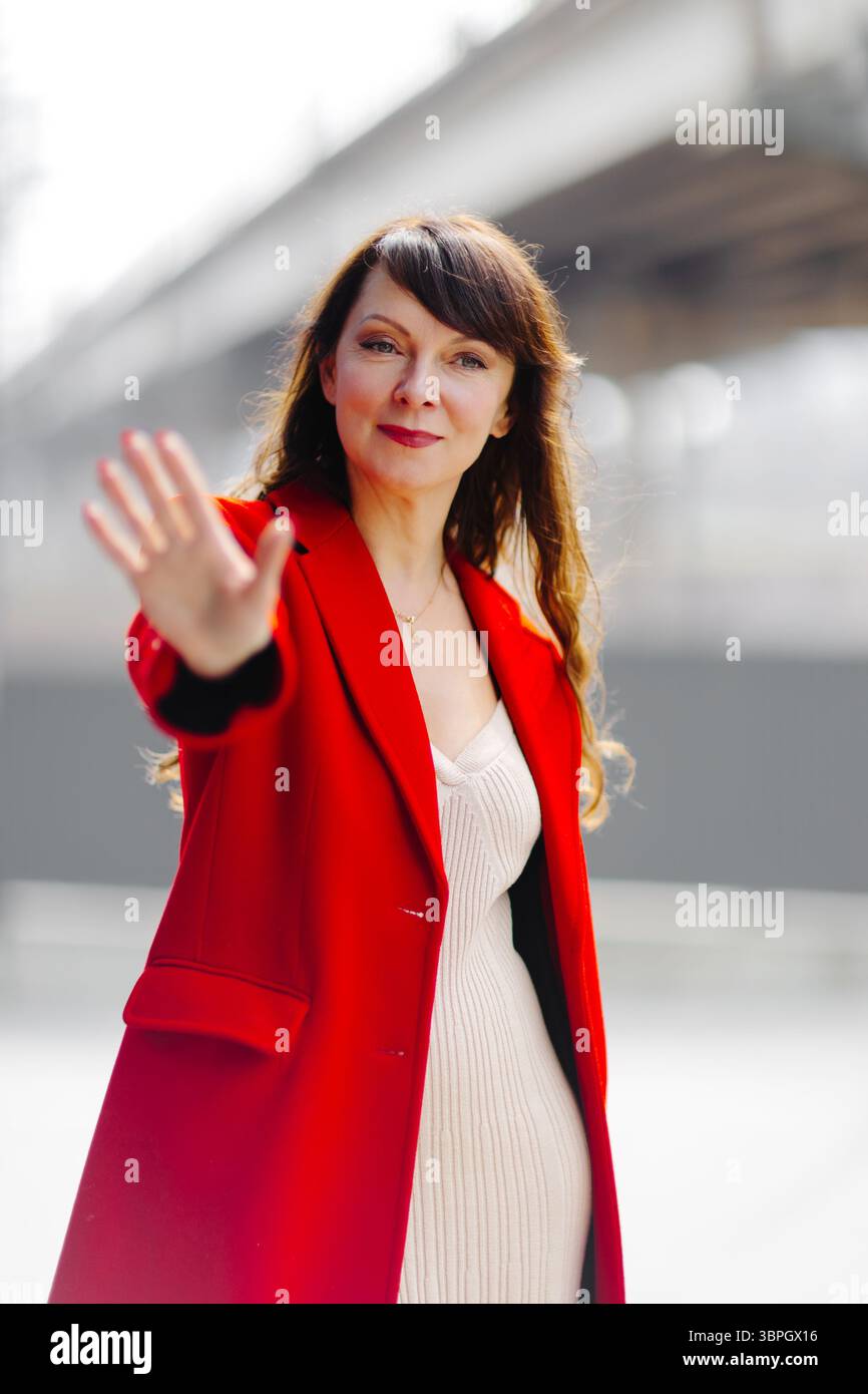 Geschäftsfrau in rotem Mantel mit Stoppschild mit der Hand, das Selbstvertrauen und Autorität in einer beruflichen Umgebung ausdrückt Stockfoto