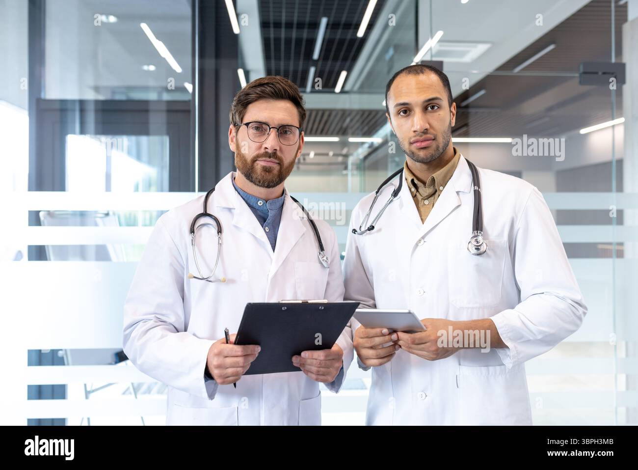 Zwei männliche Ärzte stehen zusammen, einer hält ein Klemmbrett und der andere ein Tablet, in einer modernen medizinischen Umgebung. Stockfoto