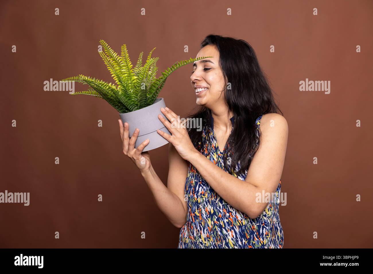 Porträt einer fröhlichen indianerin mit fröhlichem Lächeln, die die Topfpflanze hält und ihre Blätter bewundert. Glückliches weibliches Model steht vor braunem Hintergrund und begeistert mit der Freude an der Gartenarbeit. Stockfoto