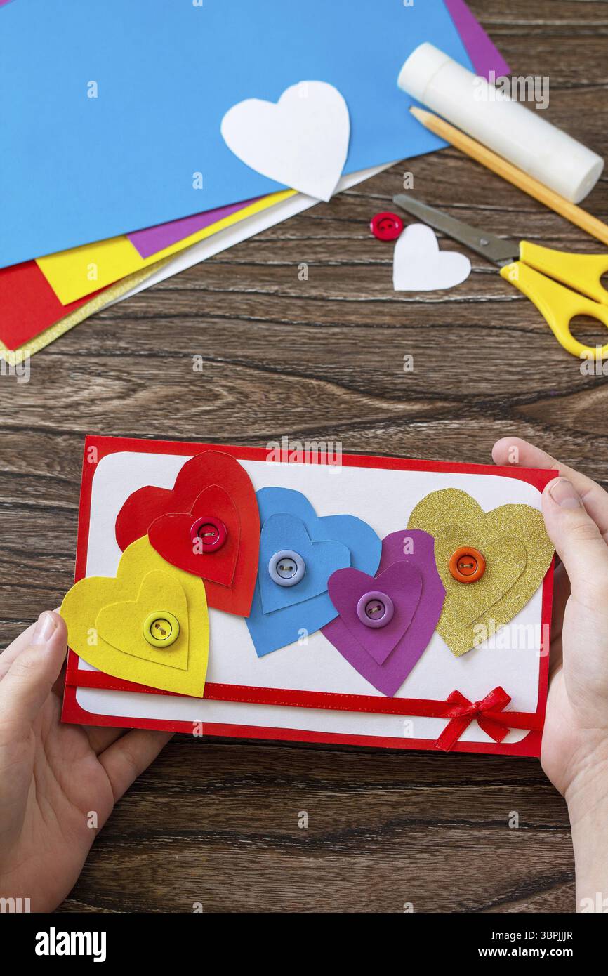 Kind hält Grußkarte mit einem Herzen Valentinstag, Tag Mutter oder Geburtstagsgeschenk auf Holztisch. Kinderkunstprojekt Kunsthandwerk für Kinder. Stockfoto
