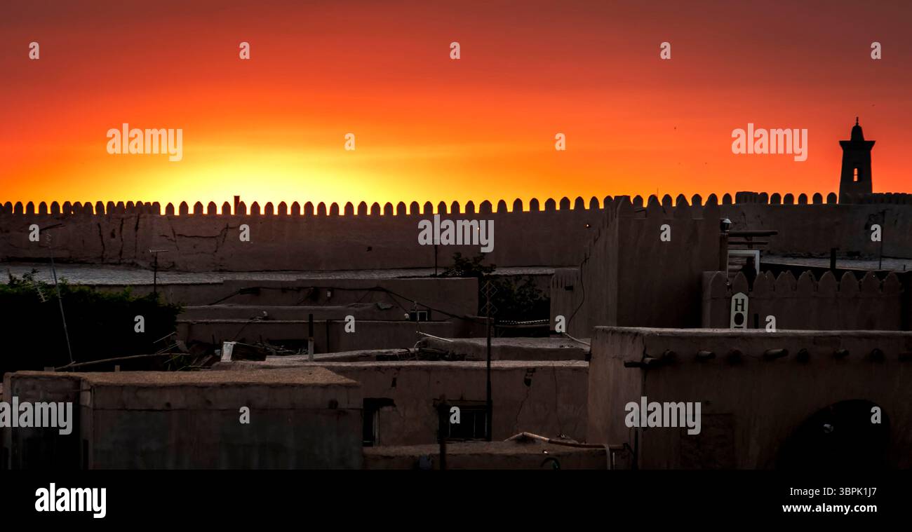 Ein atemberaubender Sonnenuntergang über einem traditionellen marokkanischen Stadtbild, mit einer Silhouette einer befestigten Mauer und einem Minarett vor einem leuchtenden orangefarbenen Himmel. Stockfoto