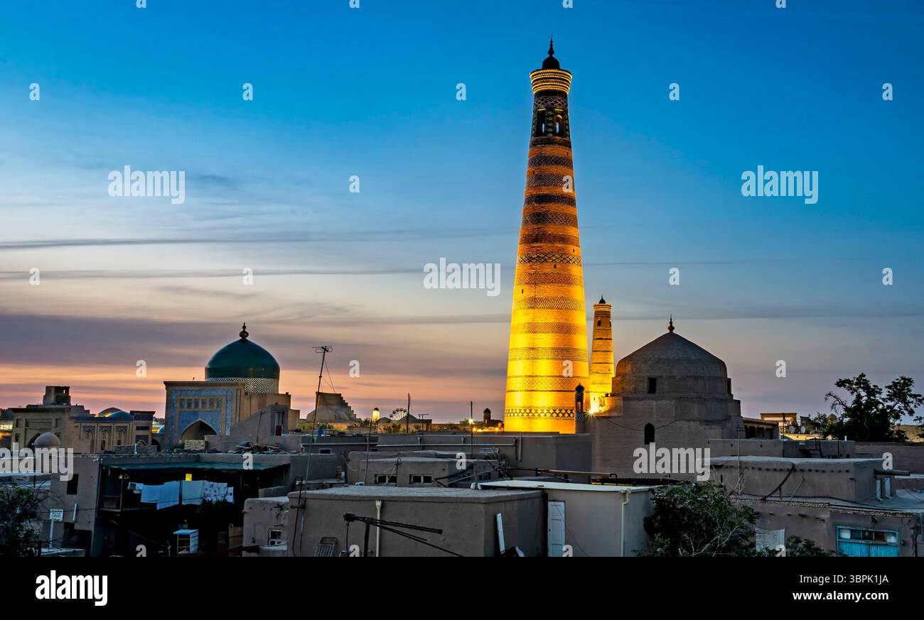 Ein atemberaubender Blick auf ein historisches Minarett, das in der Abenddämmerung beleuchtet wird, umgeben von traditioneller Architektur. Der Himmel wechselt von blau zu orange und erzeugt einen Stockfoto
