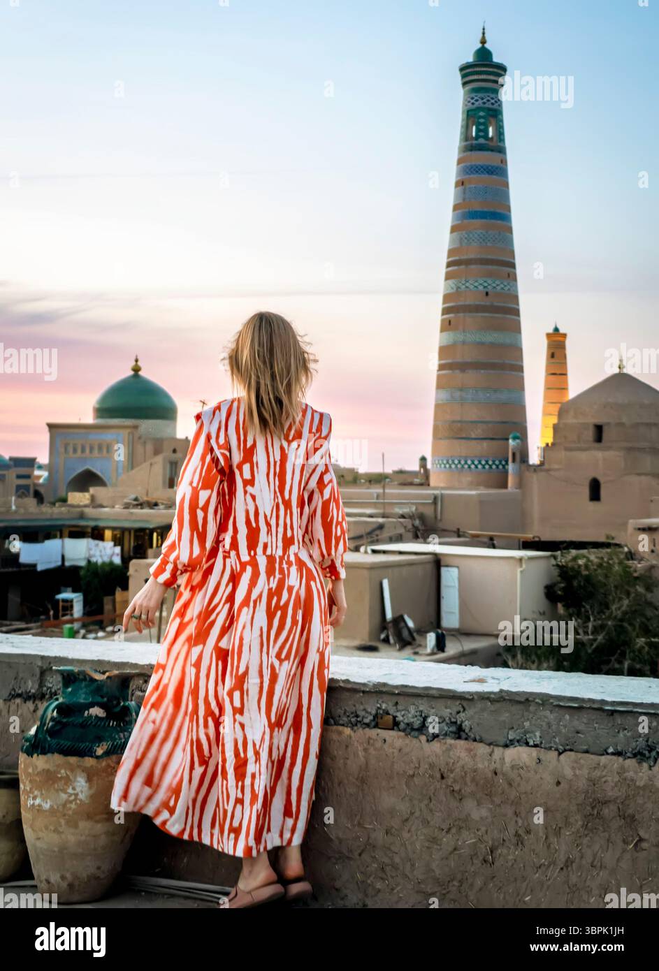 Eine Frau in einem leuchtend orange-weiß gestreiften Kleid steht auf einem Dach und bietet einen malerischen Blick auf die historische Architektur, einschließlich eines hohen Minaretts Stockfoto