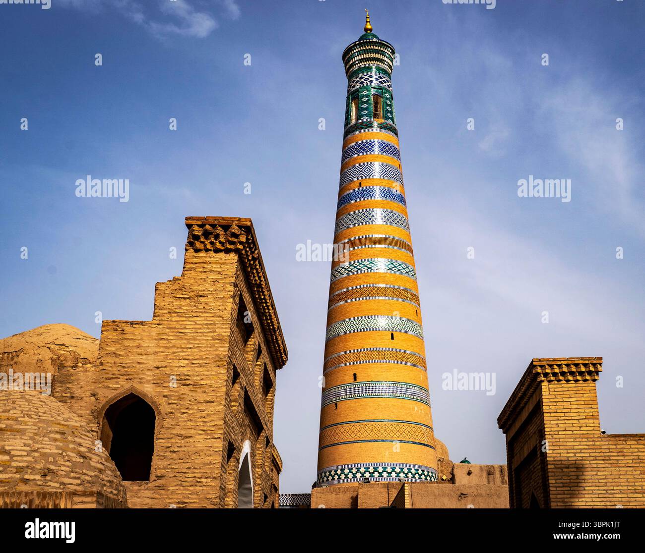 Ein hohes, farbenfrohes Minarett steht vor einem klaren blauen Himmel, umgeben von alten Backsteinbauten. Das Minarett weist komplizierte Muster auf Stockfoto