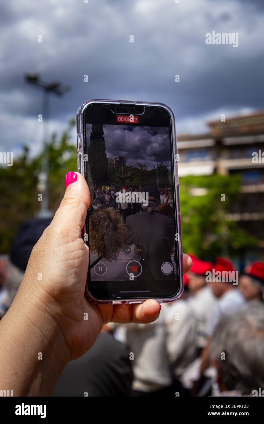 Die Hand hält ein Smartphone, während sie ein Video einer Marschkapelle aufnimmt, die während einer lebhaften Parade an einem bewölkten Tag auftritt Stockfoto