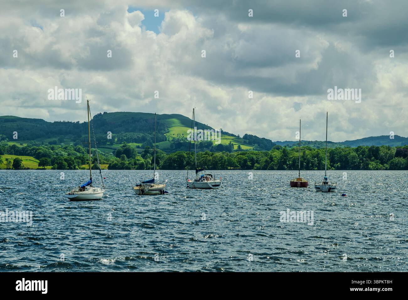 Mehrere Segelboote liegen vor dem Wind des Windermere Lake, wo an einem luftigen Sommertag weit entfernte Fjägel unter Regenwolken aufsteigen. Stockfoto