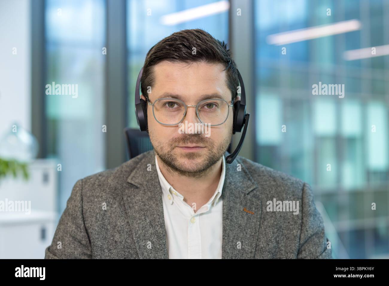 Nahporträt eines jungen seriösen männlichen Büroarbeiters und Assistenten, der mit einem Headset an seinem Arbeitsplatz sitzt und aufmerksam auf die Kamera blickt. Stockfoto