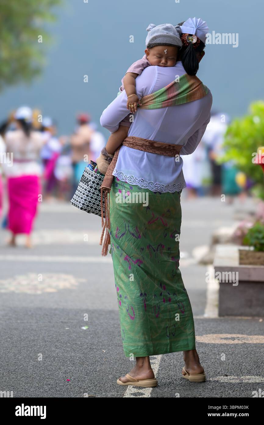 Balinesische Frau in traditioneller Kleidung, die während einer Zeremonie ein schlafendes Baby trägt, Bali, Indonesien Stockfoto